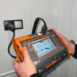 Elektryk wykonuje pomiary impedancji pętli zwarcia za pomocą miernika Sonel MPI-505, podłączonego do gniazdka elektrycznego w białej ścianie.