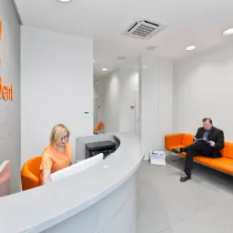 Jasne wnętrze nowoczesnej kliniki: recepcja z pracownikiem w pomarańczowej bluzce, pacjent czytający na pomarańczowej sofie, logo 'acusdent' na ścianie z teksturą.