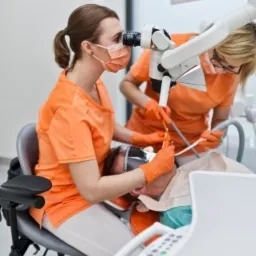 Dwaj stomatolodzy w pomarańczowych uniformach przeprowadzają zabieg na pacjencie pod mikroskopem Leica, z widocznym zdjęciem rentgenowskim w tle.