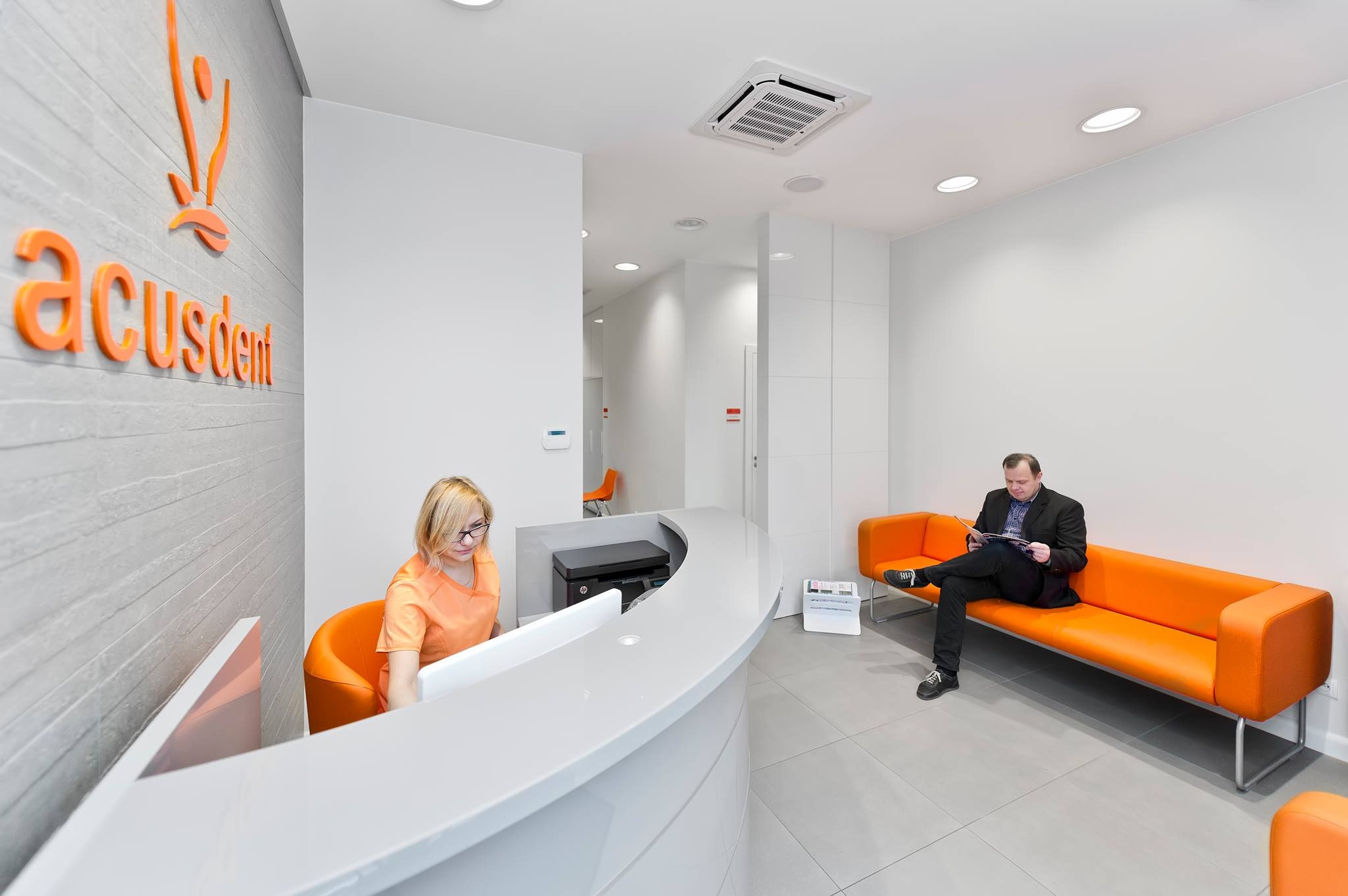 Jasne wnętrze nowoczesnej kliniki: recepcja z pracownikiem w pomarańczowej bluzce, pacjent czytający na pomarańczowej sofie, logo 'acusdent' na ścianie z teksturą.