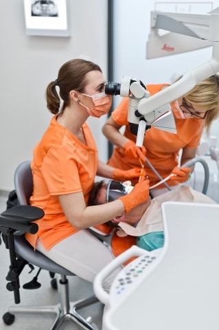 Dwaj stomatolodzy w pomarańczowych uniformach przeprowadzają zabieg na pacjencie pod mikroskopem Leica, z widocznym zdjęciem rentgenowskim w tle.