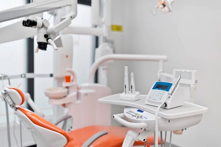 Nowoczesny gabinet stomatologiczny z pomarańczowym fotelem, mikroskopem zabiegowym i panelem sterowania urządzeniami.
