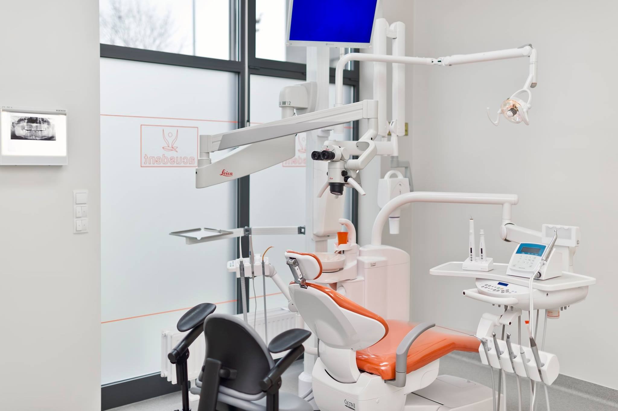 Nowoczesny gabinet stomatologiczny z pomarańczowym fotelem, mikroskopem Leica, lampą zabiegową i panoramicznym zdjęciem rentgenowskim na ścianie.
