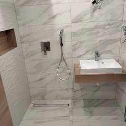 Hańderek Wojciech - Wykończona łazienka z białymi płytkami imitującymi marmur, kabina prysznicowa typu walk-in z odpływem liniowym, umywalka nablatowa na drewnianym blacie, bateria podtynkowa i deszczownica, widoczne...