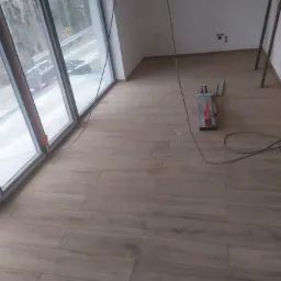 Ułożona podłoga z płytek imitujących drewno w jasnym pomieszczeniu z dużym oknem, widoczny przecinak do płytek i wystające kable.