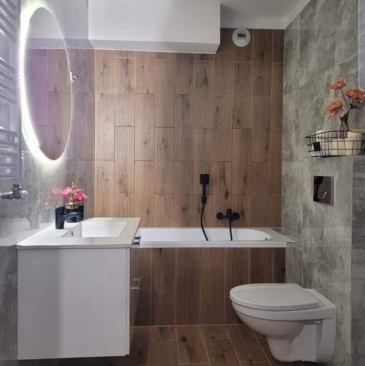 Nowoczesna łazienka w Krakowie z drewnopodobnymi płytkami na ścianach, białym zestawem umywalkowym i wanną z czarną armaturą, oświetlonym lustrem i szarymi płytkami imitującymi beton.