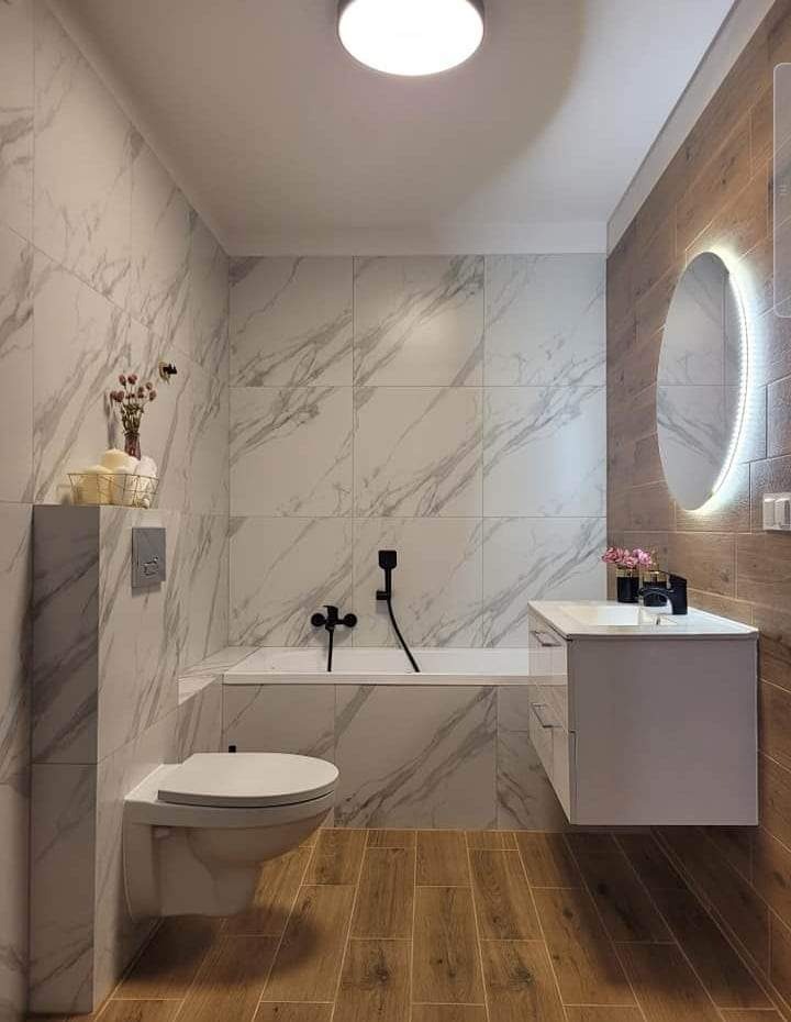 Nowoczesna łazienka z płytkami imitującymi marmur na ścianach, drewnopodobną podłogą, owalnym lustrem z podświetleniem LED, czarną armaturą i wiszącą toaletą.