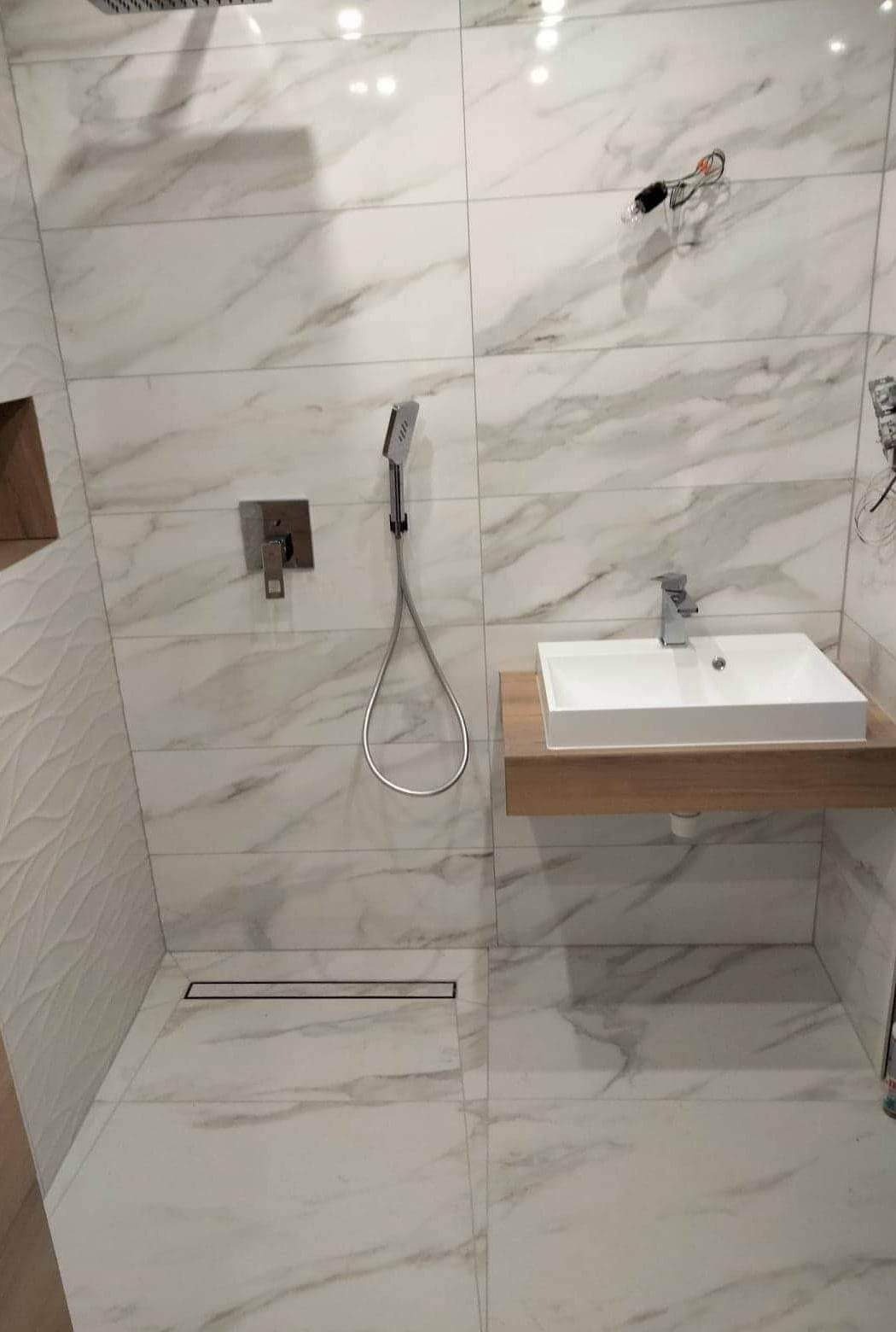 Nowoczesna łazienka z białymi płytkami imitującymi marmur, kabina prysznicowa typu walk-in z odpływem liniowym, umywalka nablatowa na drewnianym blacie, bateria podtynkowa i deszczownica.