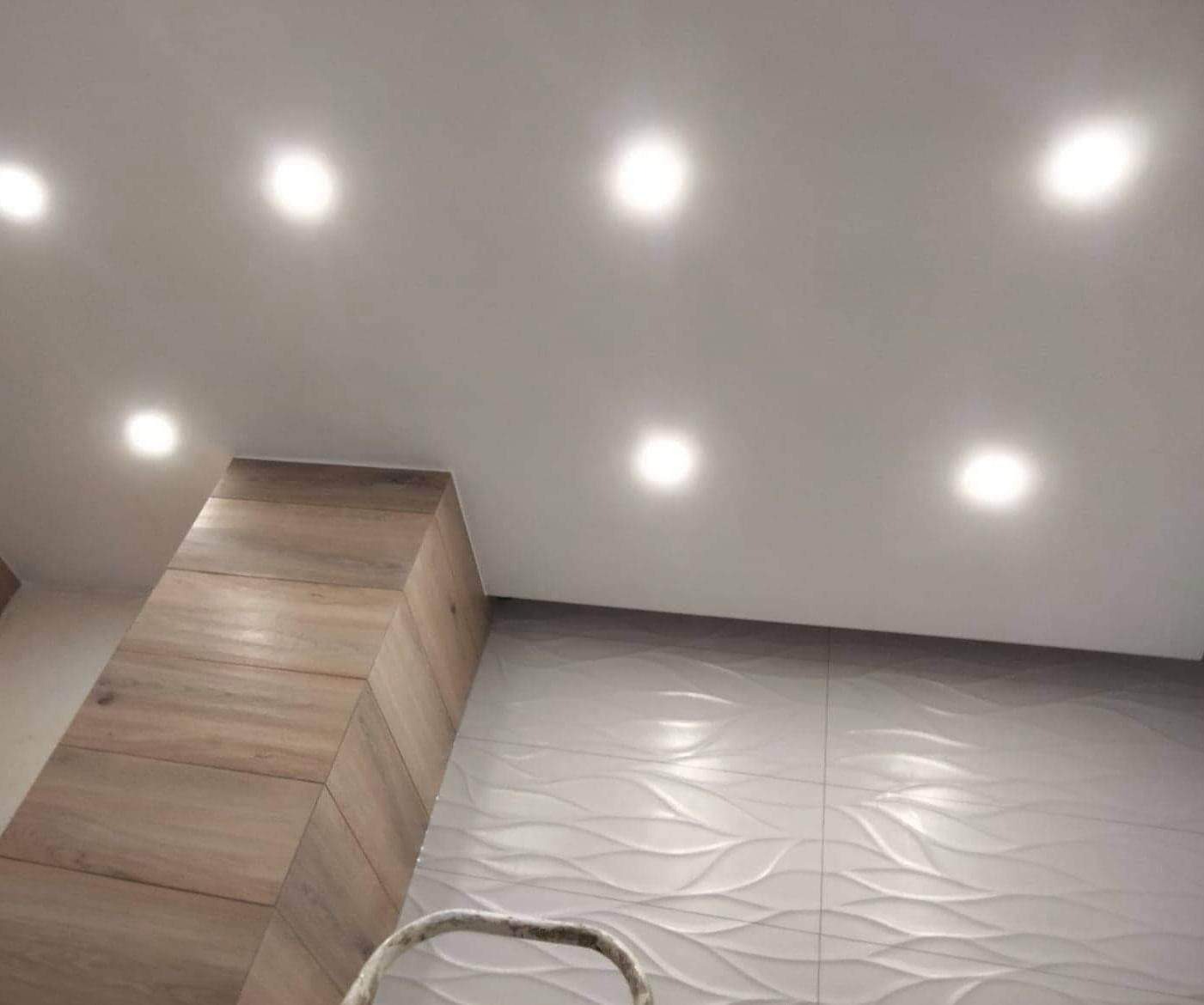 Nowoczesna łazienka po remoncie z podwieszanym sufitem i oświetleniem punktowym, dekoracyjnymi płytkami ściennymi z motywem fal oraz obudową pionu z płytek imitujących drewno.