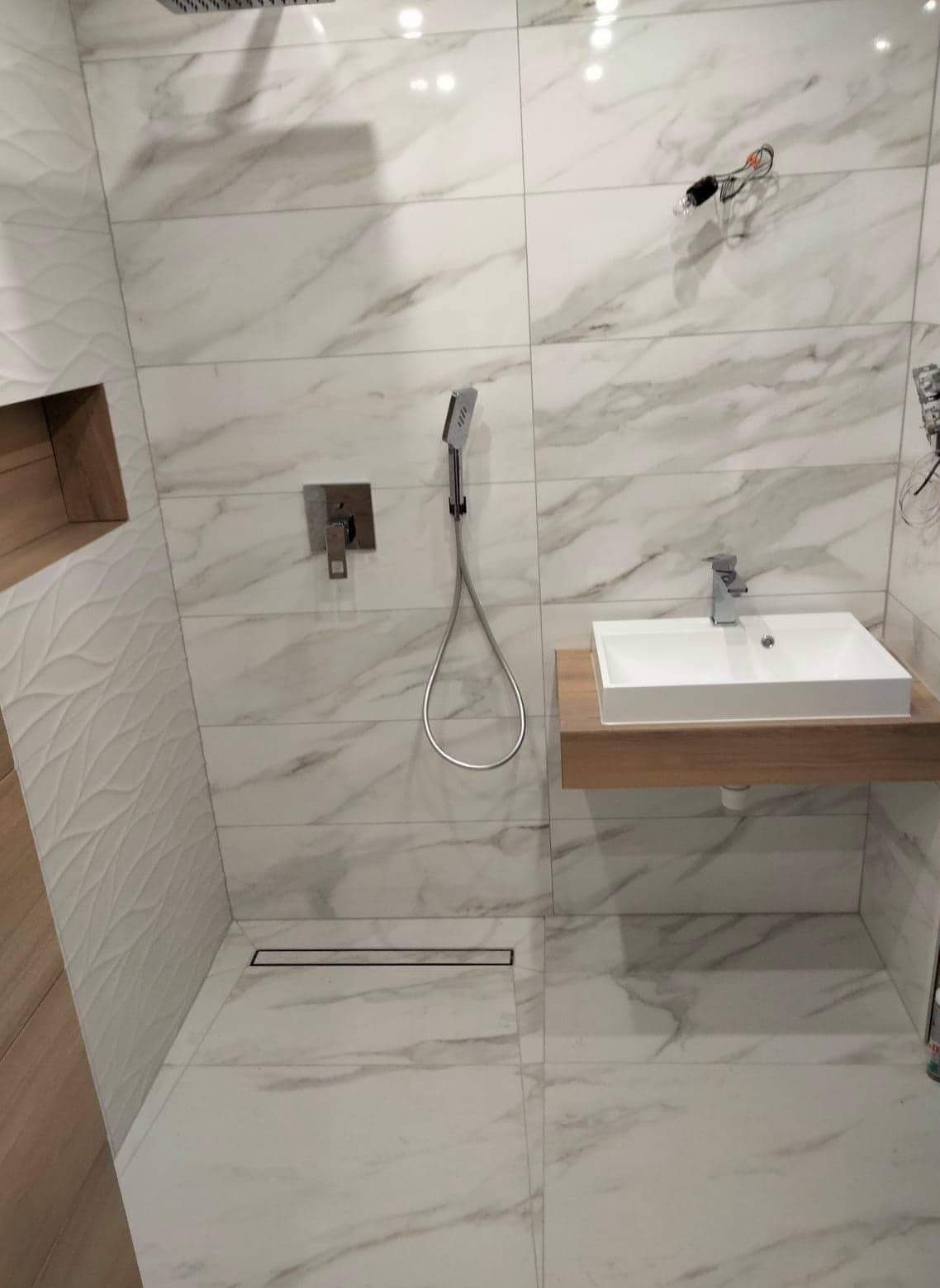 Wykończona łazienka z białymi płytkami imitującymi marmur, kabina prysznicowa typu walk-in z odpływem liniowym, umywalka nablatowa na drewnianym blacie, bateria podtynkowa i deszczownica, widoczne...