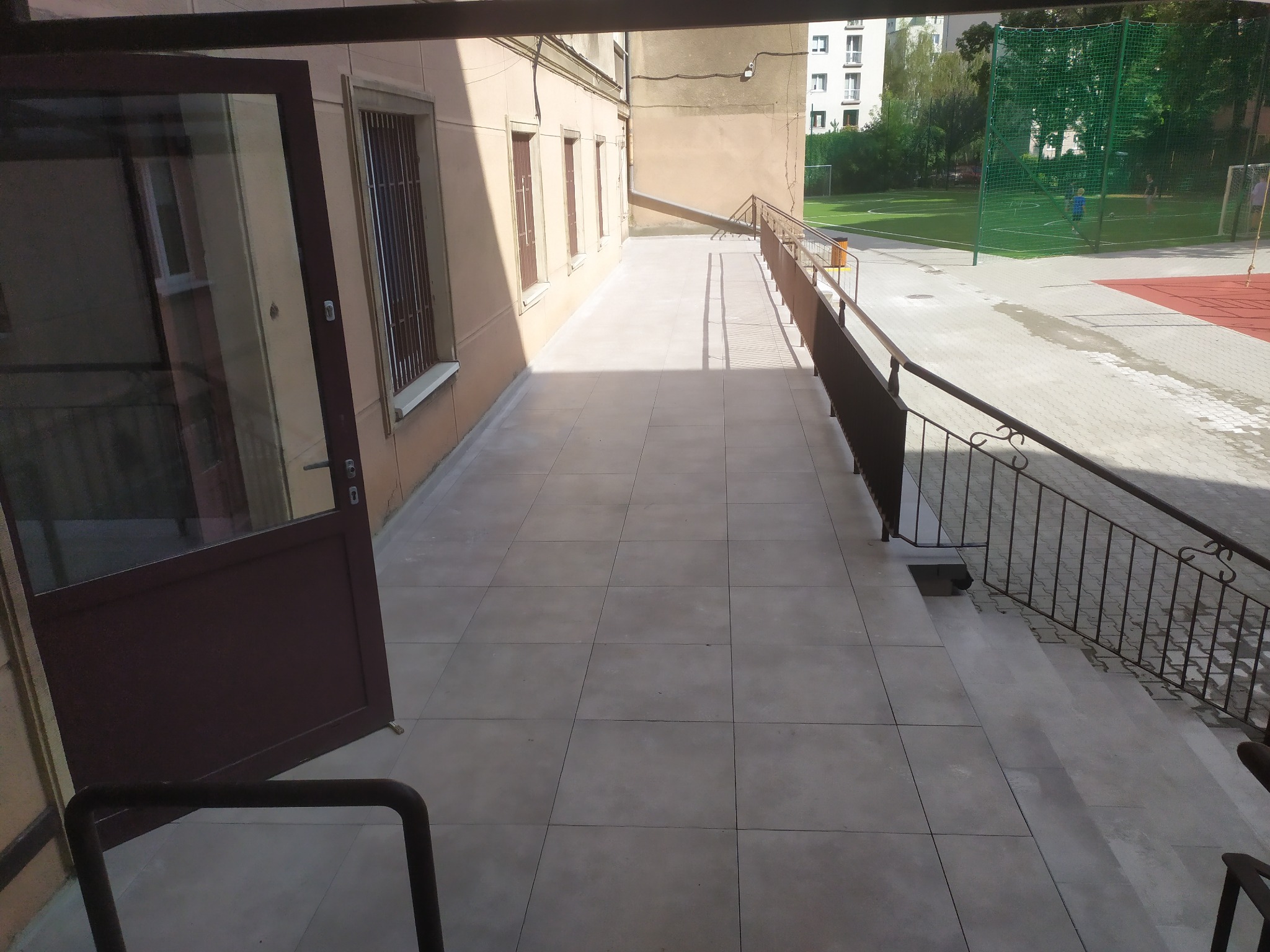Wyłożony szarymi płytkami taras przed budynkiem z otwartymi drzwiami, widok na boisko sportowe w tle.