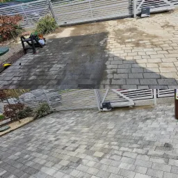 Częściowo wyczyszczona kostka brukowa przed posesją z bramą wjazdową, widoczne różnice w odcieniach szarości, ślady wilgoci i szczotka do czyszczenia.