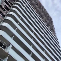 Wysoki budynek mieszkalny z widocznymi rusztowaniami na górnych piętrach, sfotografowany pod kątem od dołu, ukazujący liczne balkony i pochmurne niebo w tle.