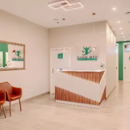 Nowoczesne wnętrze centrum medycznego Hand-Med w Radomiu: recepcja z logo firmy, dwie skórzane fotele w poczekalni, lustro z odbiciem informacji dla pacjentów oraz korytarz z numerowanymi drzwiami...