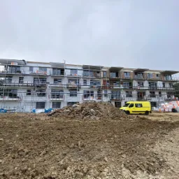 Wielopiętrowy budynek w trakcie prac elewacyjnych, z widocznymi rusztowaniami, oknami i fragmentami ocieplenia, na pierwszym planie hałda ziemi, a w tle żółty samochód dostawczy i stosy materiałów...