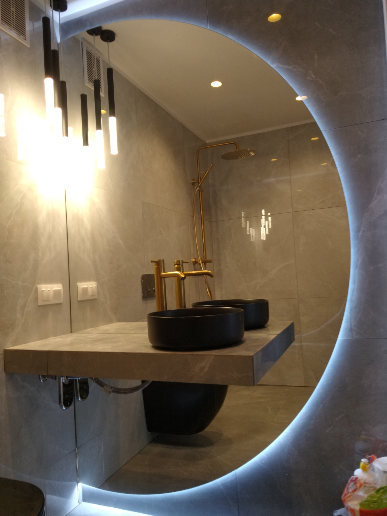 Nowoczesna łazienka z dwoma czarnymi umywalkami nablatowymi, złotą armaturą, podświetlanym lustrem w kształcie koła i wiszącymi lampami.