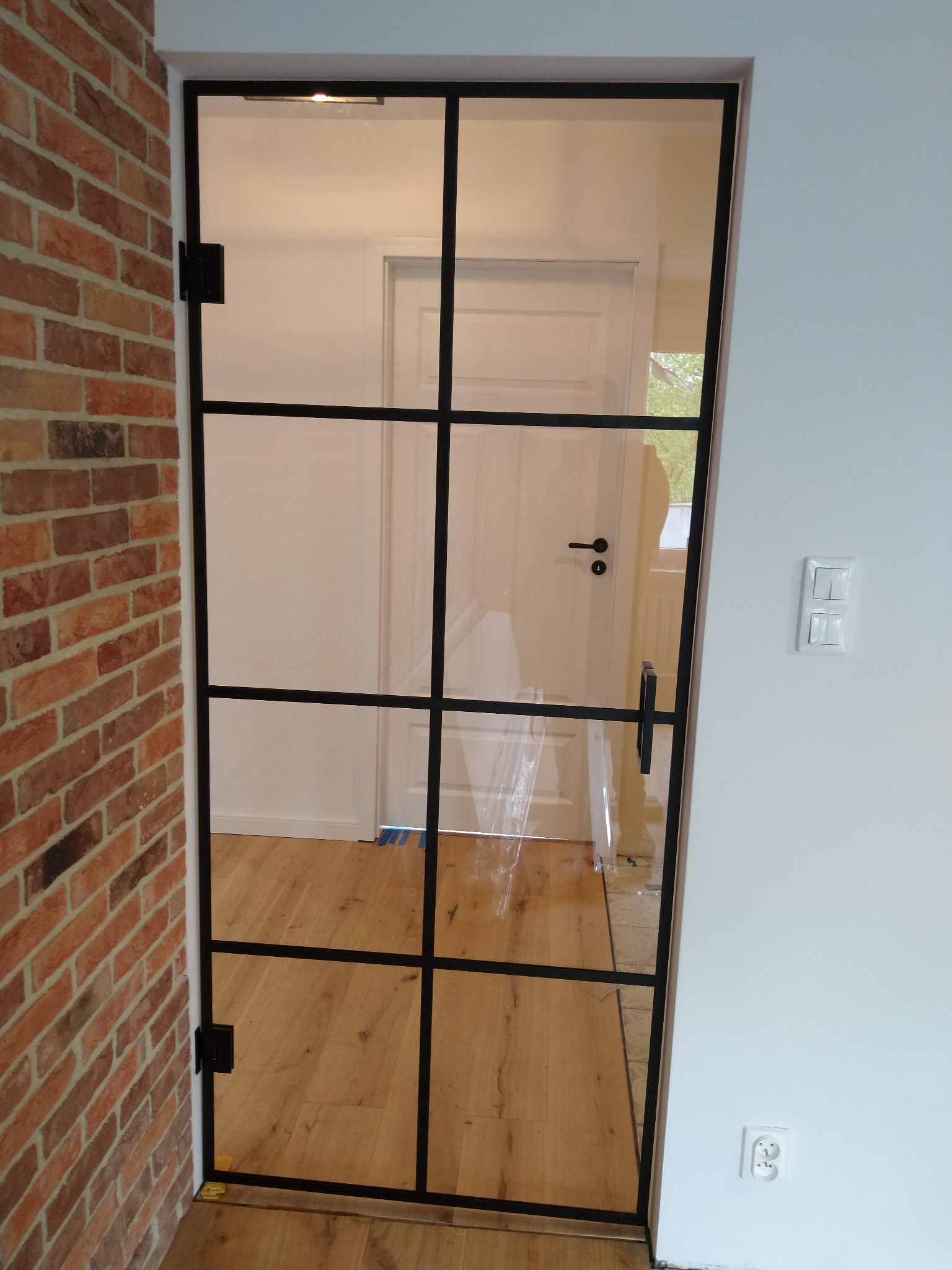 Szklane drzwi wewnętrzne z czarną ramą i podziałami, zamontowane między ścianą z cegły a białą ścianą z włącznikiem światła i gniazdkiem.