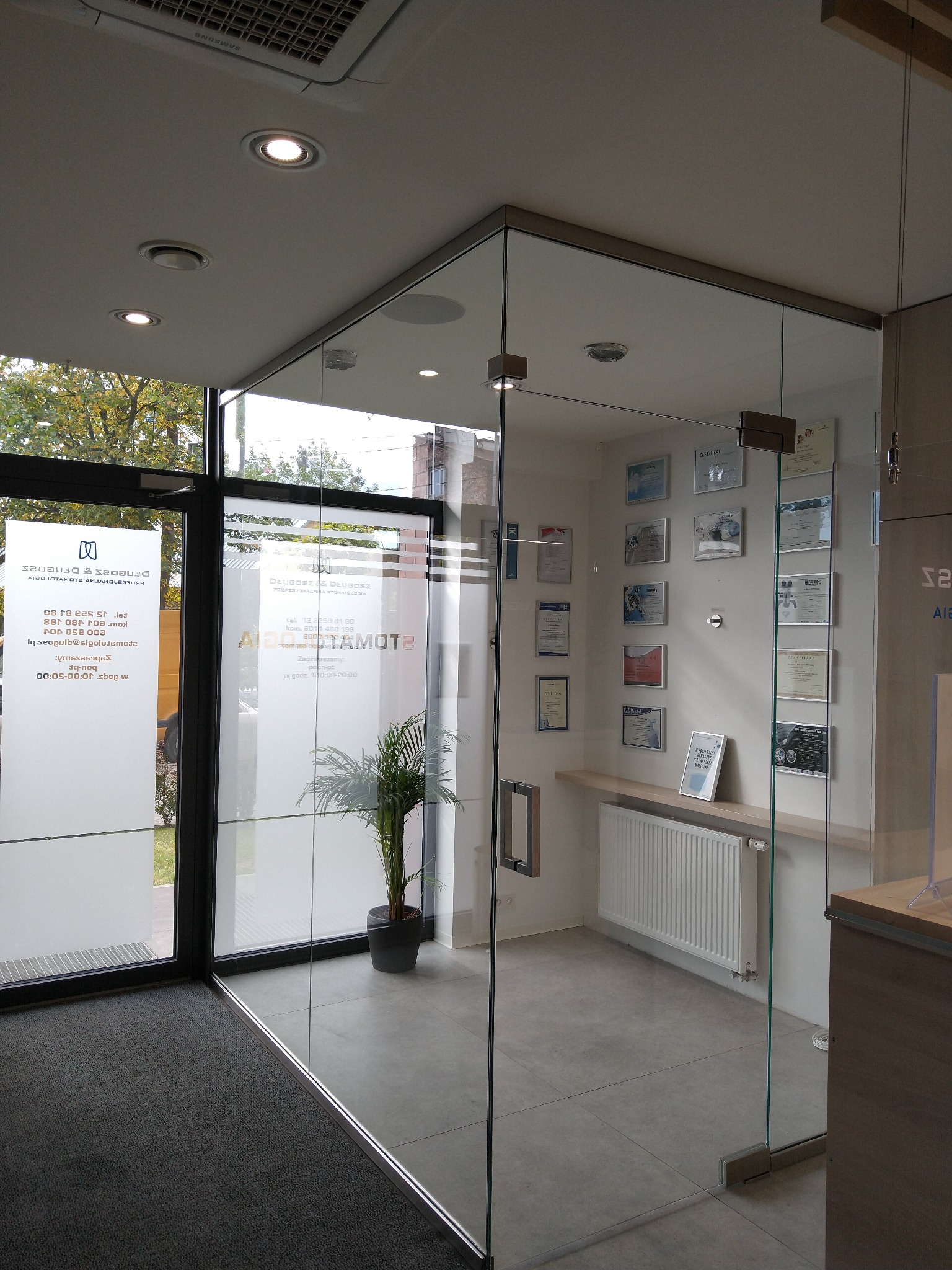Szklana ścianka działowa z drzwiami w biurze, widoczne certyfikaty na ścianie i roślina w doniczce, szklane drzwi wejściowe z naklejkami informacyjnymi.