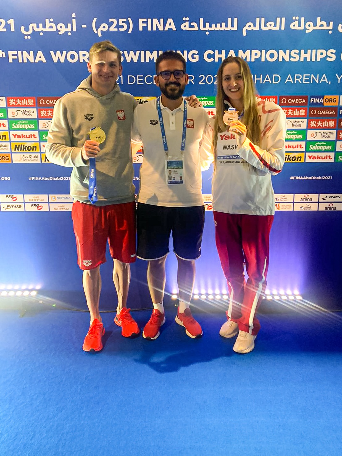 Trener pływania z dwoma medalistami Mistrzostw Świata w Abu Dhabi 2021, Radosławem Kawęckim i Katarzyną Wasick, stojący na tle baneru mistrzostw.