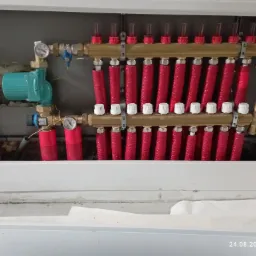 Rozdzielacz ogrzewania podłogowego z pompą obiegową w szafce, widoczne czerwone rury i manometry.