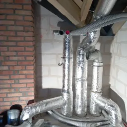 Instalacja rekuperacji w surowym stanie w Jarosławiu: widoczne srebrne rury izolowane folią aluminiową, szare rury karbowane na podłodze, ceglana ściana w tle.