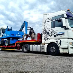 Niebieska maszyna Fuchs MHL 331 z długim ramieniem, prawdopodobnie koparka, transportowana na czerwonej, niskopodwoziowej naczepie ciągniętej przez białą ciężarówkę MAN z czarnym motywem skorpiona...