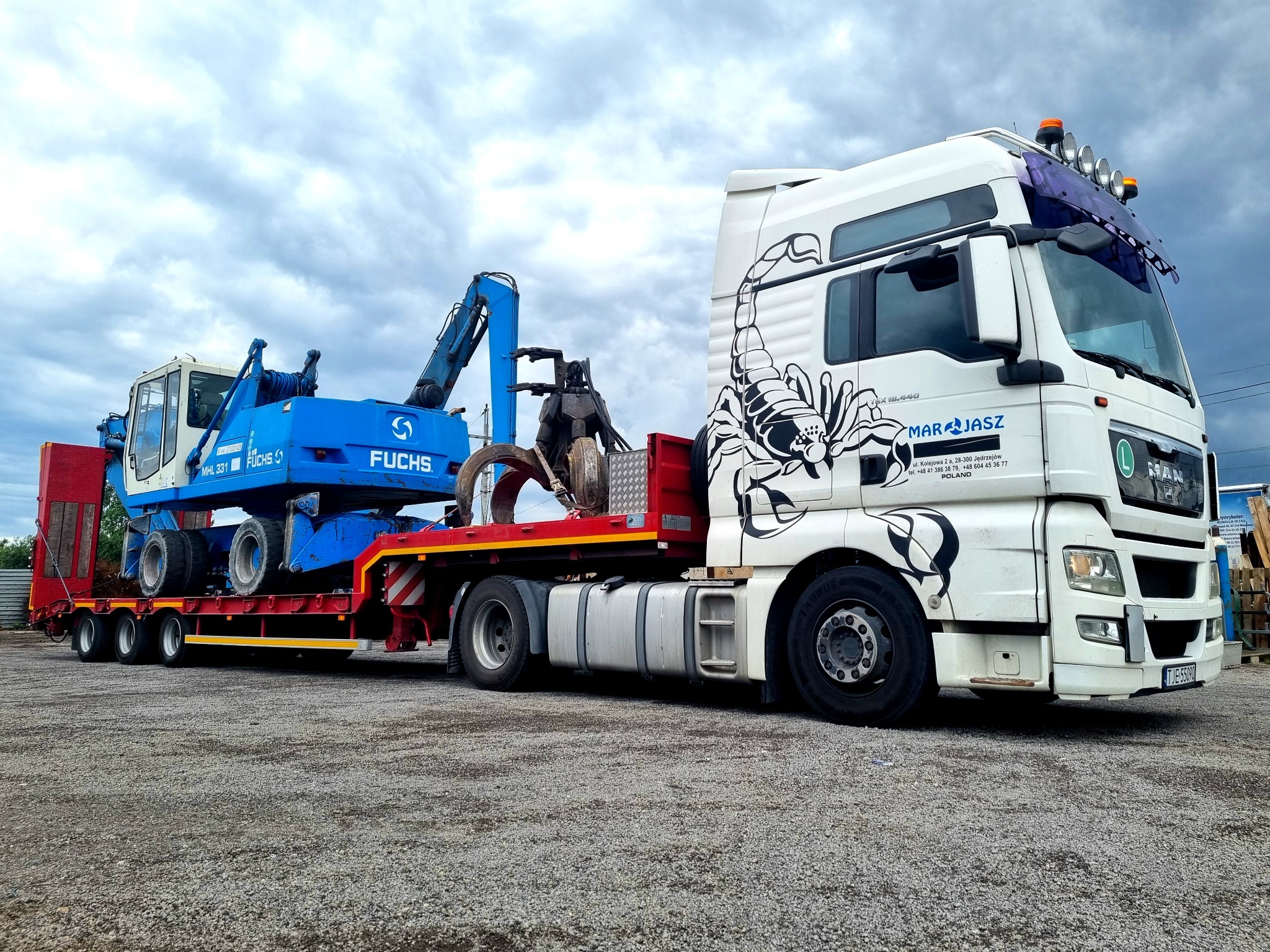 Niebieska maszyna Fuchs MHL 331 z długim ramieniem, prawdopodobnie koparka, transportowana na czerwonej, niskopodwoziowej naczepie ciągniętej przez białą ciężarówkę MAN z czarnym motywem skorpiona...
