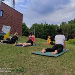 Grupa kobiet ćwiczy na matach do ćwiczeń na trawie przed budynkiem z cegły, wykonując pozycję kobry, z widocznym banerem reklamowym w tle.