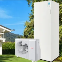 Biała pompa ciepła powietrze-woda, składająca się z jednostki zewnętrznej i wewnętrznej, zainstalowana na trawniku przed domem z zielonymi krzewami i błękitnym niebem w tle.