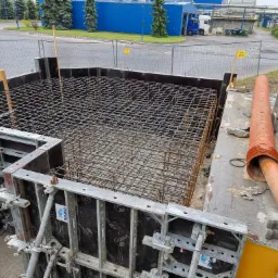 Zbrojenie fundamentu w szalunku z widocznymi elementami mocującymi, rurą wentylacyjną i drabiną w tle.