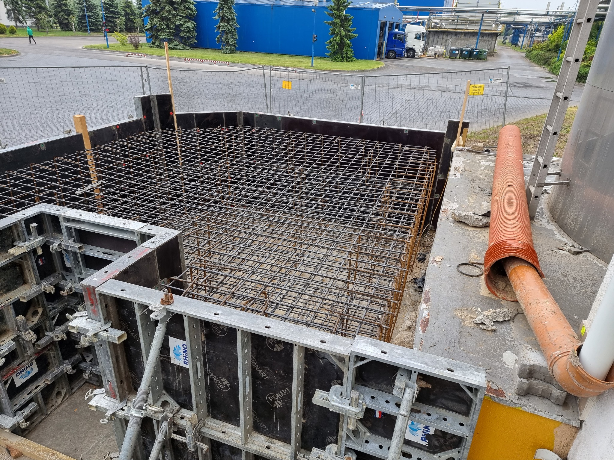Zbrojenie fundamentu w szalunku z widocznymi elementami mocującymi, rurą wentylacyjną i drabiną w tle.