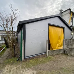 Szara budowla z paneli, prawdopodobnie garaż lub budynek gospodarczy, z żółtą plandeką w otworze drzwiowym, na tle pochmurnego nieba i częściowo wykończonego domu w trakcie budowy.