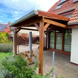 Drewniana pergola z brązowym dachem przylegająca do domu z czerwonym dachem i panelami słonecznymi, widok z ogrodu z zielonymi krzewami i szarymi słupkami.