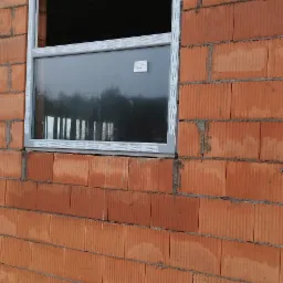 Montaż okna PCV w surowej ścianie z czerwonej cegły ceramicznej, widoczne drewniane elementy szalunku u dołu ściany.