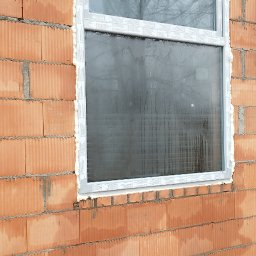 Jag-Woj - Nowo zamontowane okno PCV w surowej ścianie z czerwonej cegły, uszczelnione pianką montażową, widoczne krople wody na szybie.