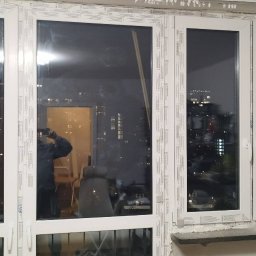 Jag-Woj - Montaż okna PCV z widoczną pianką montażową, nocny widok miasta odbijający się w szybach, zabezpieczające taśmy na ramach, w tle widoczna drabina i sylwetka osoby robiącej zdjęcie.
