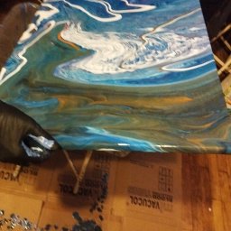 Jag-Woj - Abstrakcyjny obraz z efektem płynącej żywicy epoksydowej w odcieniach błękitu, bieli i złota, zabezpieczany czarną rękawicą, leżący na kartonowym podłożu z napisami 'VACUCOL BLOOD COLLECTION TUBES'.