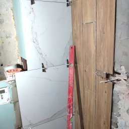 Jag-Woj - Ściana łazienki w trakcie układania płytek imitujących drewno oraz płytek marmurowych, widoczny poziomica i krzyżyki dystansowe.