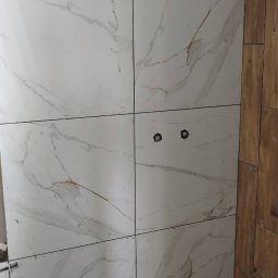 Jag-Woj - Ściana w trakcie układania płytek imitujących marmur, z krzyżykami dystansowymi, widoczne otwory na gniazdka, poziomica laserowa na wiaderku, częściowo wykończona ściana z paneli drewnopodobnych obok.