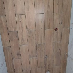 Jag-Woj - Ściana łazienki wyłożona drewnopodobnymi płytkami ceramicznymi w trakcie montażu, widoczne przyłącza instalacji wodnej, na podłodze płytka z przyssawkami do przenoszenia.