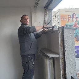 Jag-Woj - Pracownik demontuje obudowę okna balkonowego za pomocą młotka i dłuta, stojąc na drabinie; widoczny grzejnik i plakat filmowy w tle.