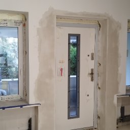 Jag-Woj - Wnętrze w trakcie prac wykończeniowych: osadzone nowe okna PCV z folią ochronną na ramach, zamontowane drzwi wejściowe z małym szybem, ściany z widocznymi śladami szpachlowania i przygotowania...