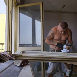 Jag-Woj - Mężczyzna z tatuażami szlifuje ramę okienną elektryczną szlifierką oscylacyjną, widoczne narzędzia i materiały przygotowane do pracy.