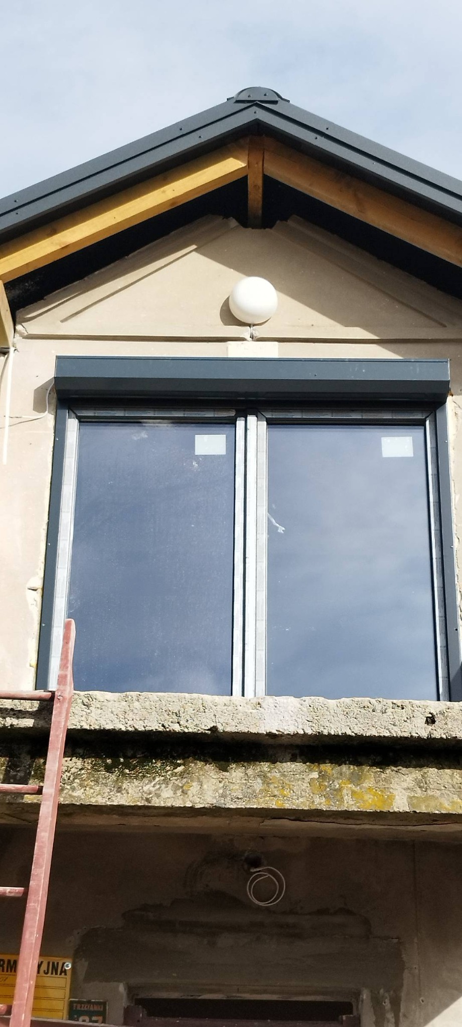 Nowe, dwuskrzydłowe okno z szarą roletą zewnętrzną zamontowane w budynku z beżową elewacją, widoczny fragment drabiny oparty o parapet.