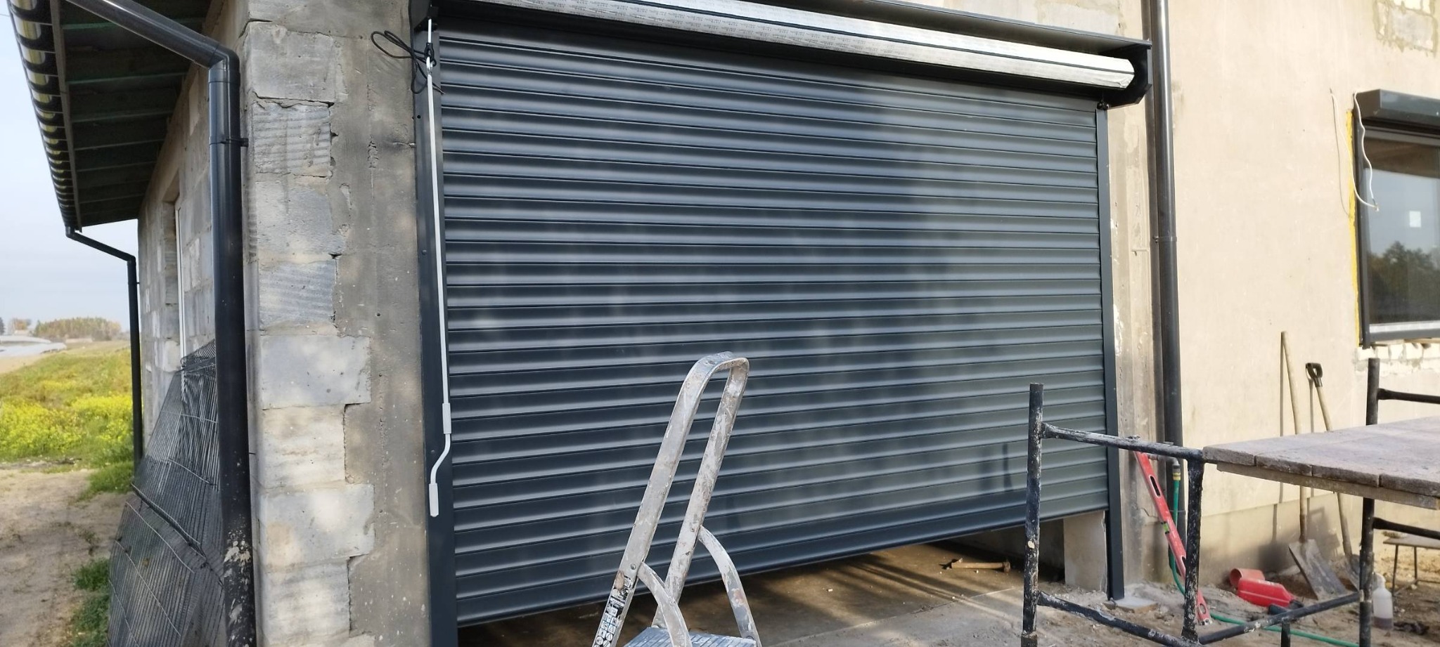 Częściowo opuszczona, szara roleta zewnętrzna na budynku, widoczne rusztowanie i drabina, budynek w trakcie prac wykończeniowych z widocznymi elementami elewacji i rynną.