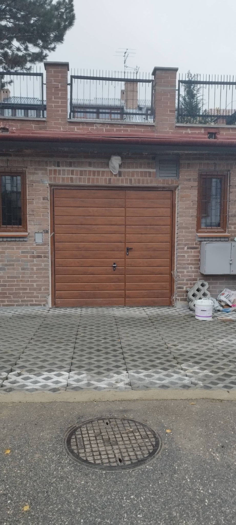 Brązowa brama garażowa segmentowa z poziomymi panelami, zamontowana w ceglanym budynku z dwoma oknami z kratami, widok na podjazd z ażurowych płyt betonowych, na ścianie biały worek zabezpieczający