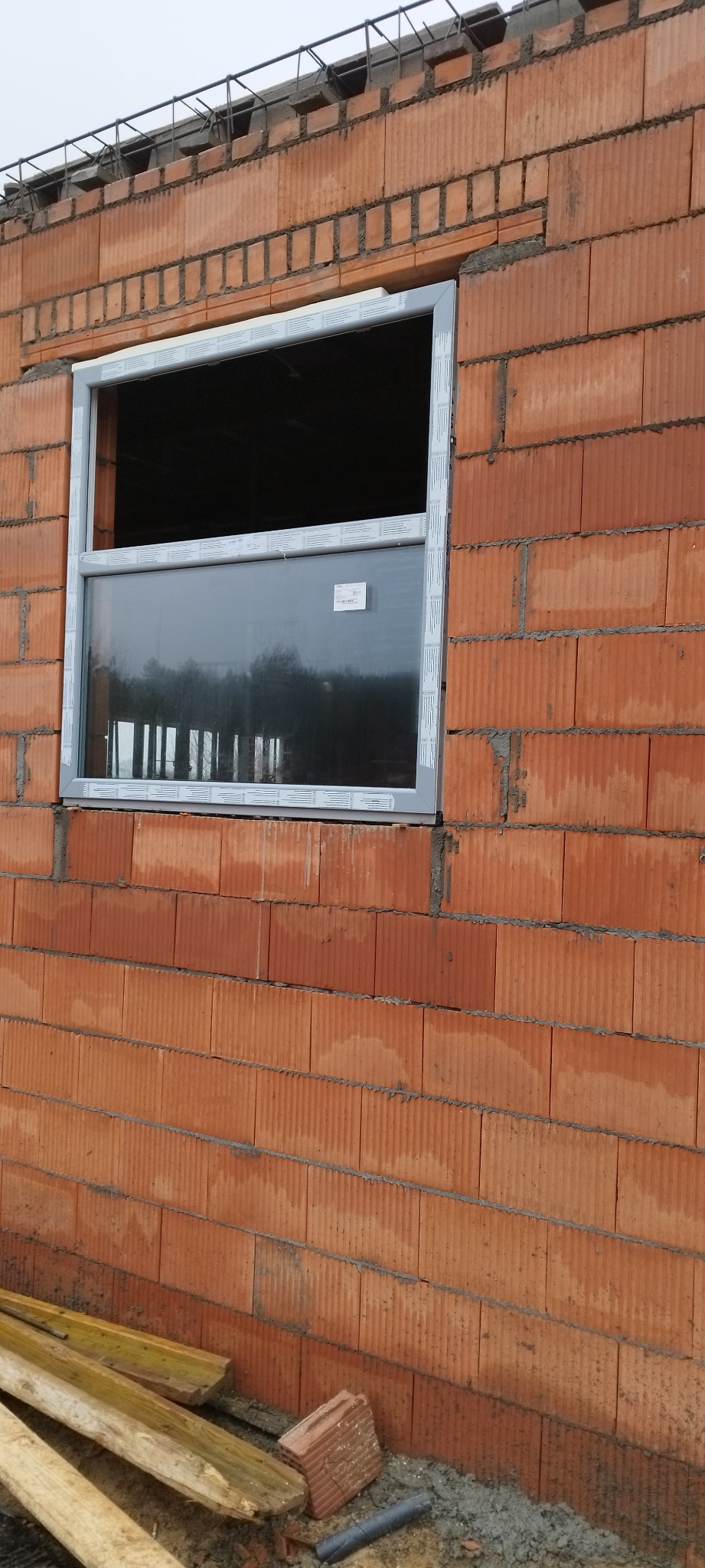 Montaż okna PCV w surowej ścianie z czerwonej cegły ceramicznej, widoczne drewniane elementy szalunku u dołu ściany.