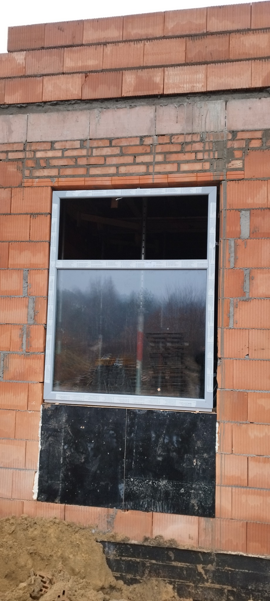 Nowe okno PCV z podwójną szybą zamontowane w surowej ścianie z czerwonej cegły, z widoczną czarną izolacją pod oknem i stertą ziemi u podstawy.