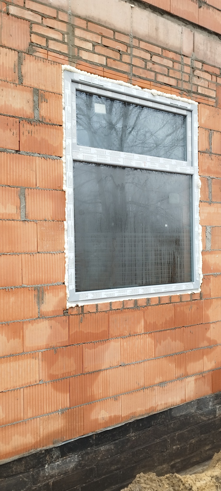 Nowo zamontowane okno PCV w surowej ścianie z czerwonej cegły, uszczelnione pianką montażową, widoczne krople wody na szybie.