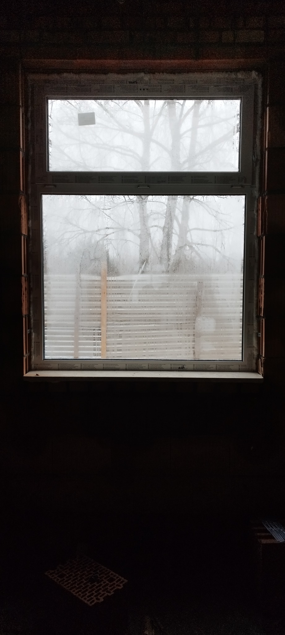 Nowe okno PCV z widokiem na zewnątrz, zamontowane w surowym murze z cegieł, widoczne ślady pianki montażowej.